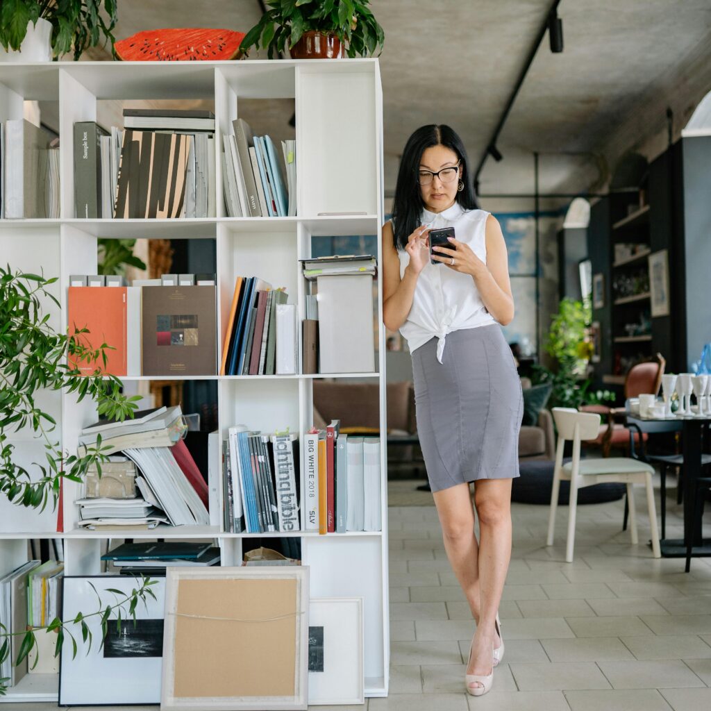 Professional woman in office wearing sleeveless shirt and gray skirt, using smartphone next to bookshelves.